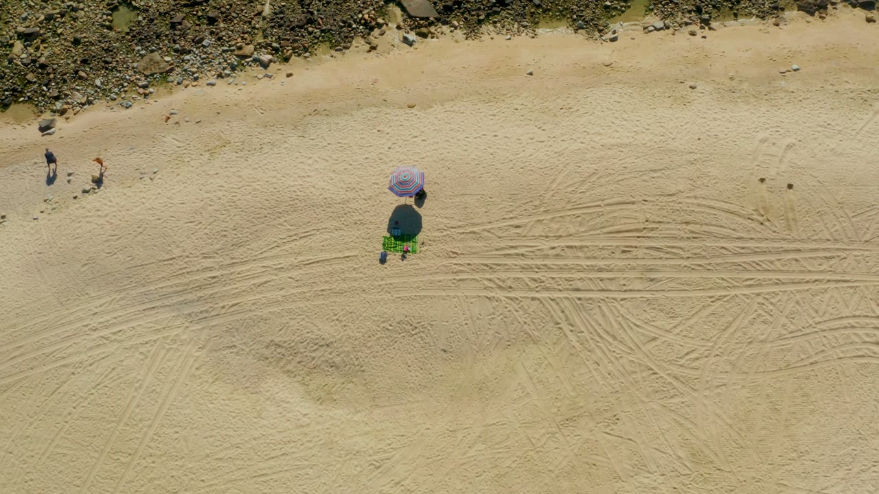 At 0:02, the Chileno Beach Los Cabos frame keeps the rocky top edge and tire tracks visible as two beachgoers move across the sand near the purple umbrella area.