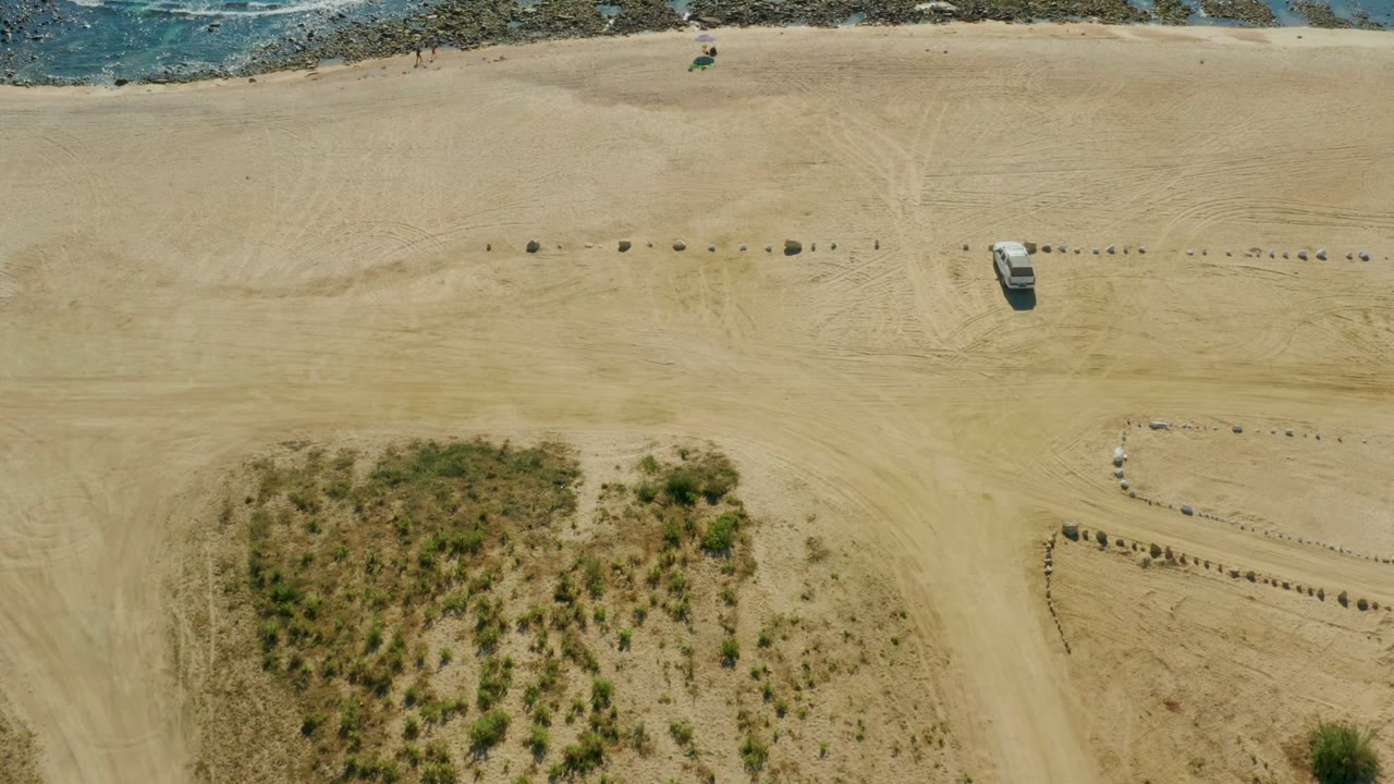 Aerial frame over Chileno Beach in Los Cabos showing pale sand, a white vehicle near the center, dune grass in the lower left, scattered rocks, and the ocean edge along the top.