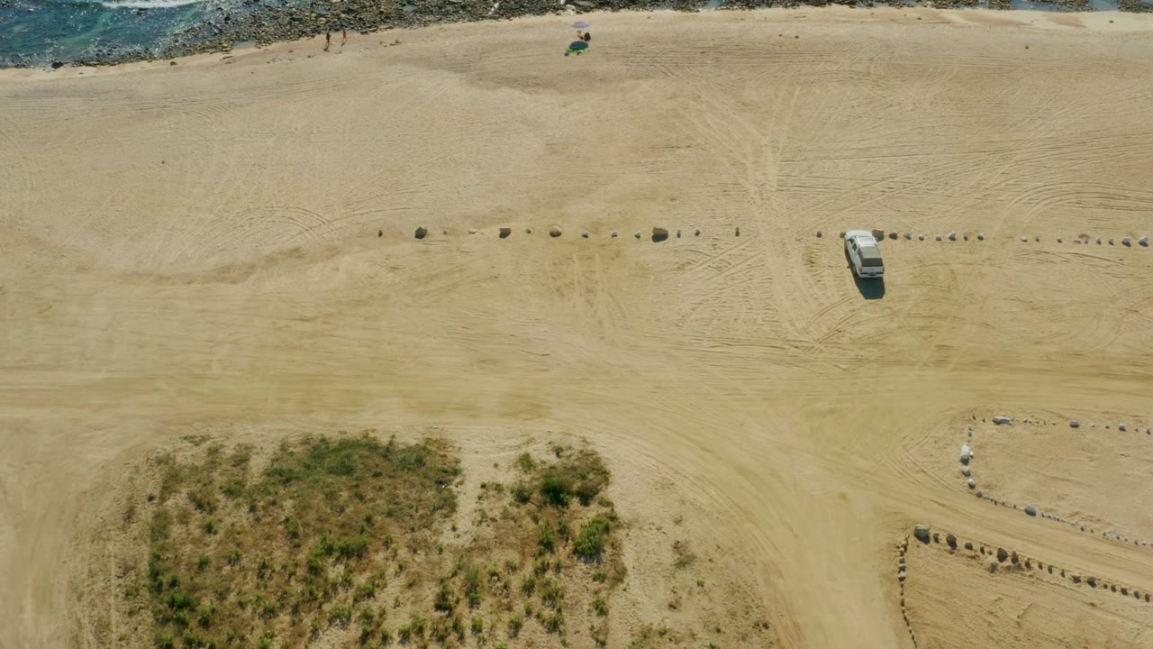 Top-down beach view with white vehicle, sand tracks, rocks, and shoreline.