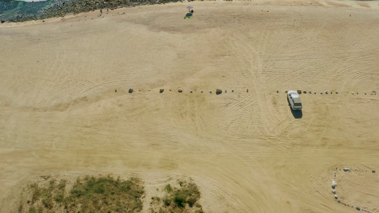 Aerial frame over Chileno Beach in Los Cabos showing a white vehicle on open sand, curved tire tracks to the right, scattered rocks, ocean at the top, and small people near the shoreline and shade tent.
