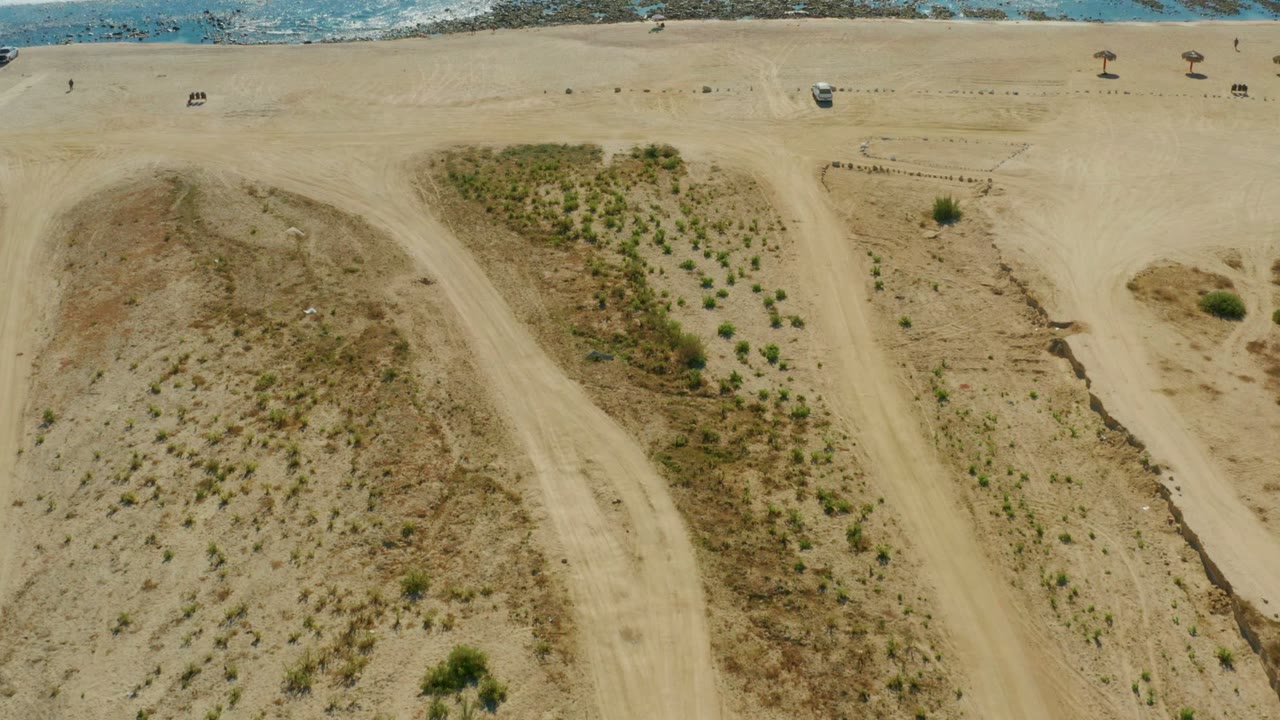 Overhead view of Chileno Beach sand dunes with sparse shrubs, pale dirt paths, and blue surf along the upper edge, with no visible people.