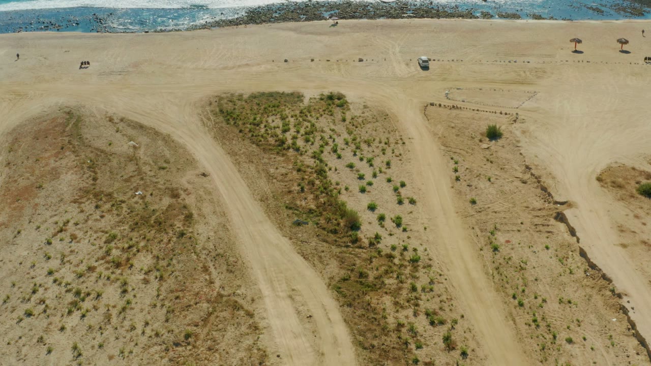 Aerial view of sandy beach with dunes, shrubs, tracks, and a strip of surf.