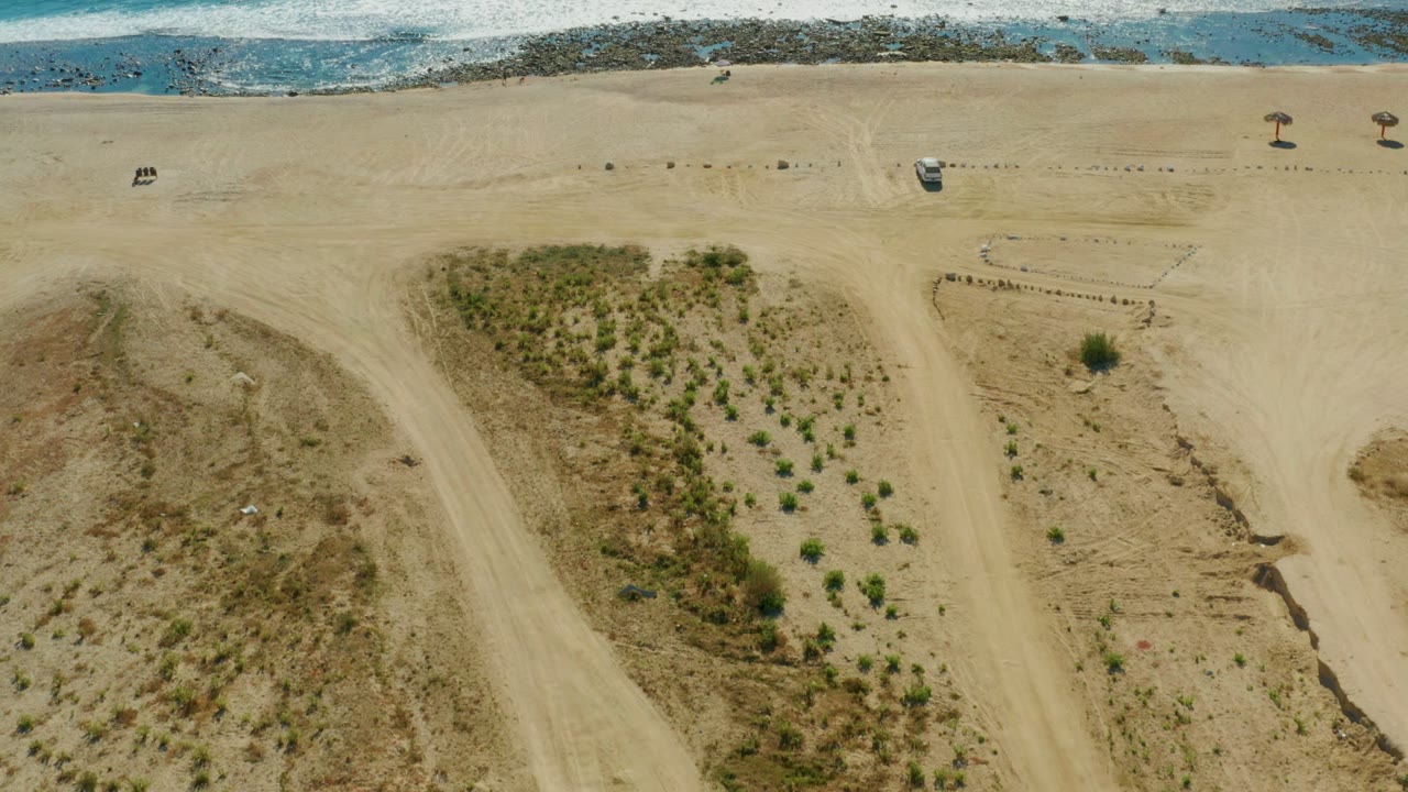 High overhead view of Chileno Beach with patterned shrub rows on sand, a parked vehicle near the shore, small shade structures, and breaking waves across the upper frame, with no visible people.