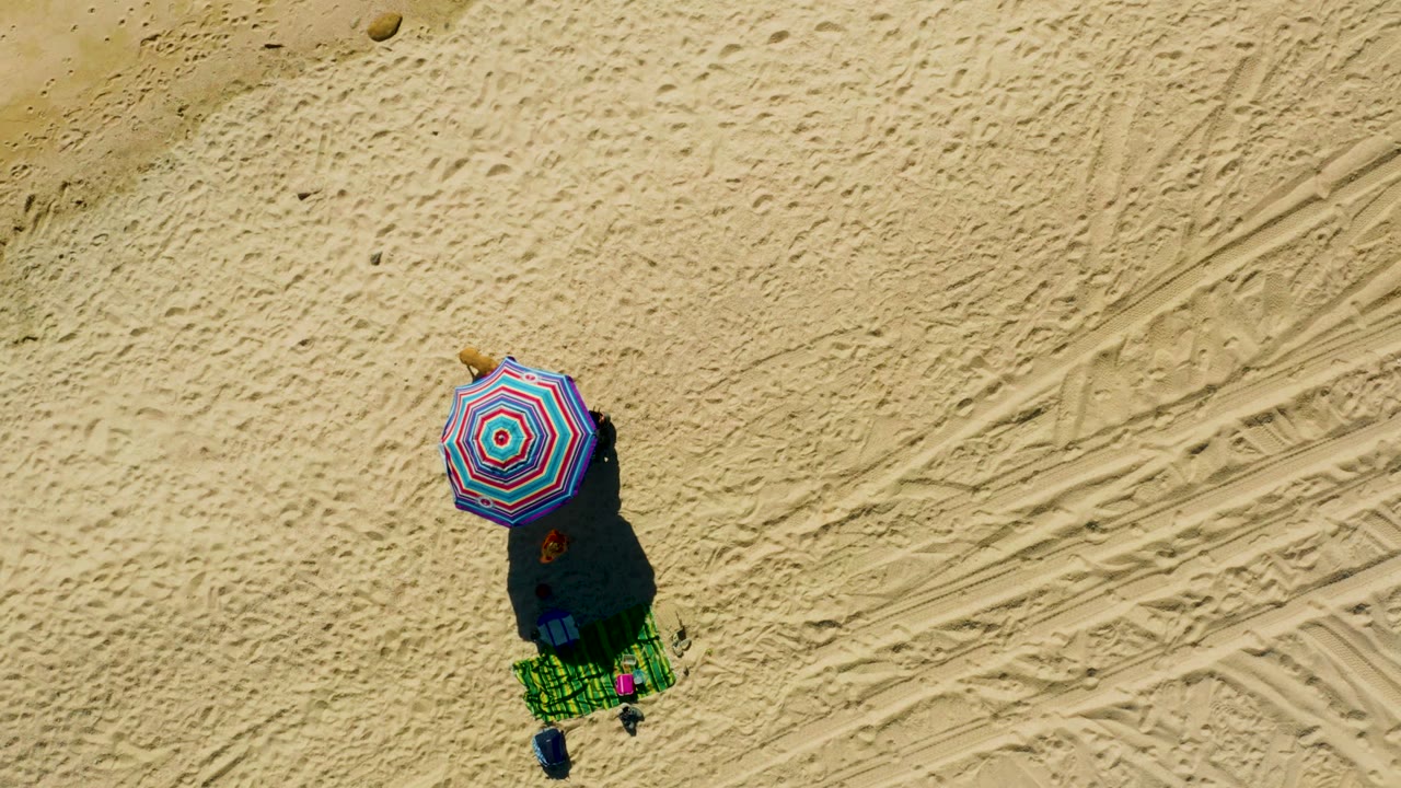 Overhead frame at Chileno Beach Los Cabos showing a red, blue, and white striped umbrella above a green towel setup on pale tracked sand, with no visible people in this moment.