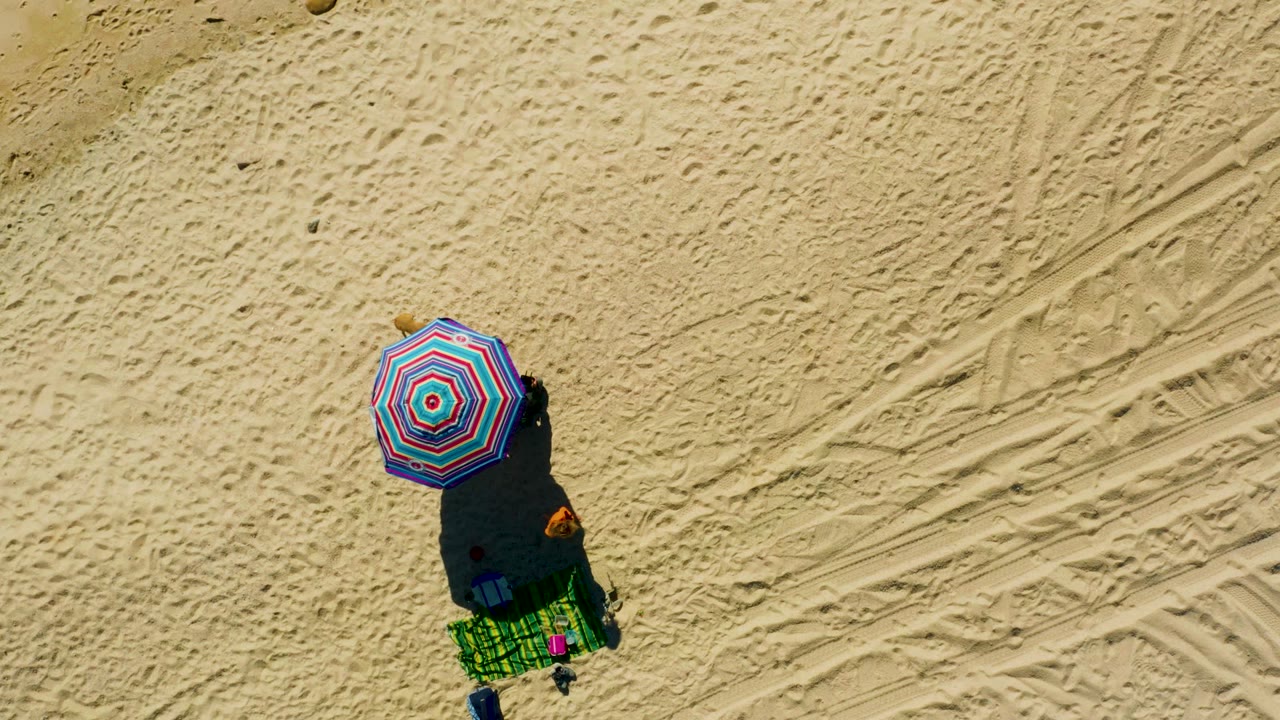 Top-down beach umbrella, towel setup, bag, and shadow on tracked sand.