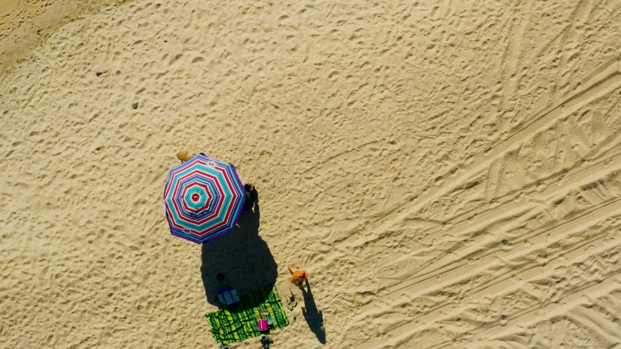 Overhead Chileno Beach Los Cabos frame showing the striped umbrella, green towel setup, blue bag, and a person in an orange top standing to the right on the sand.