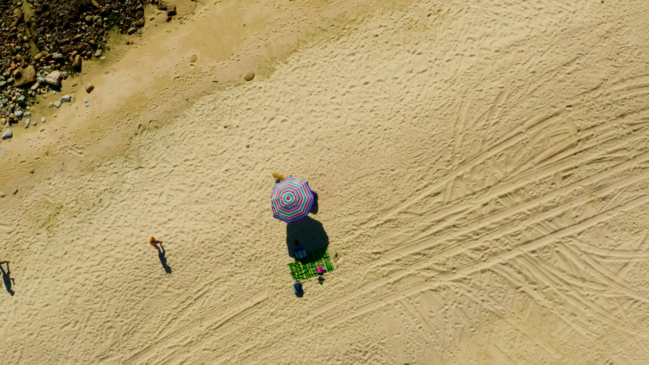 Overhead view of Chileno Beach in Los Cabos with a striped umbrella on sand, a green blanket beneath it, tire tracks to the right, rocks along the upper edge, and one person walking at left.