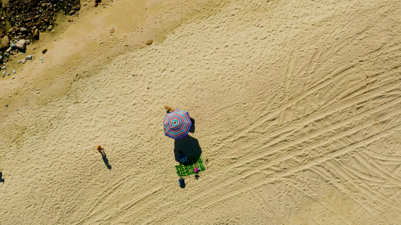Aerial sand view with colorful umbrella, green blanket, tire tracks, rocks, and a person farther from the umbrella.
