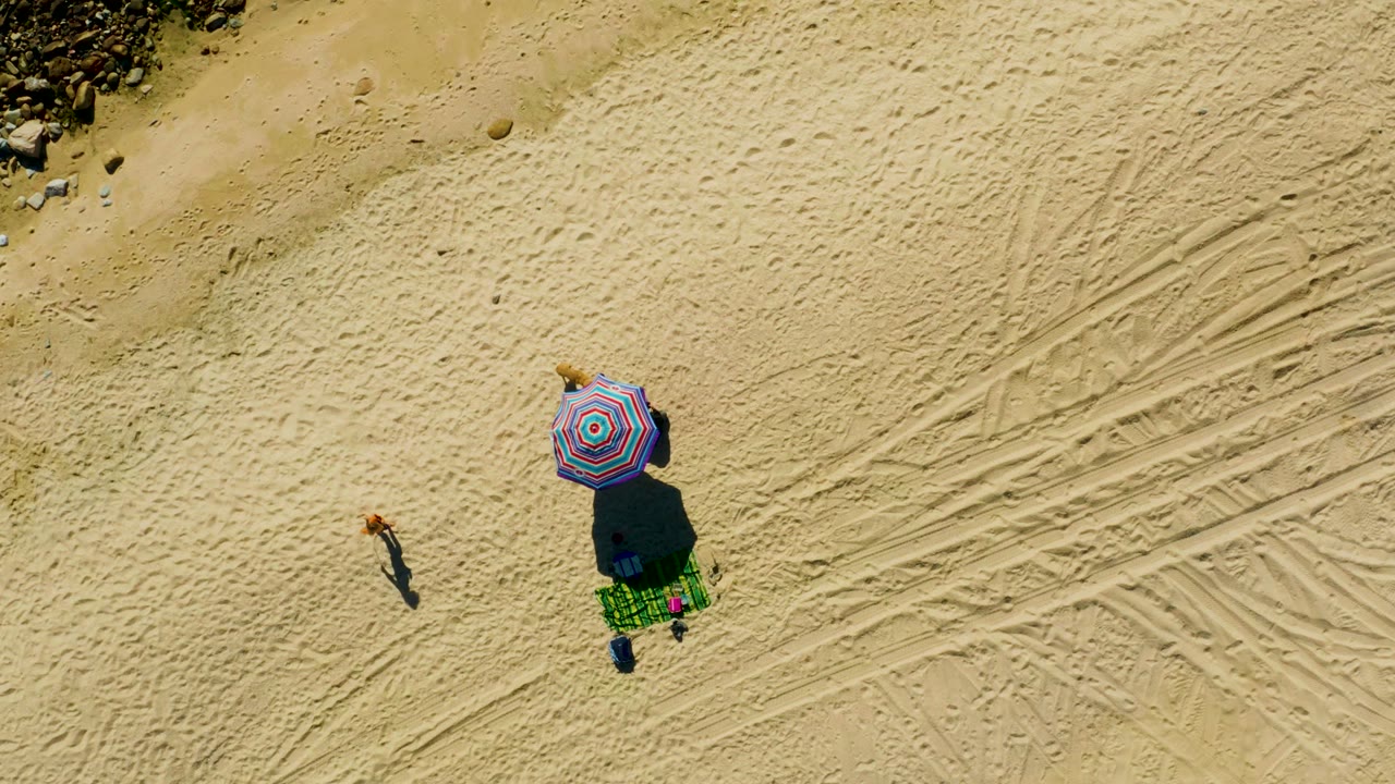 At 0:02, the overhead Chileno Beach view keeps the striped umbrella near center, the green blanket and beach items below it, one walker left of center, and tire tracks etched through the right half.