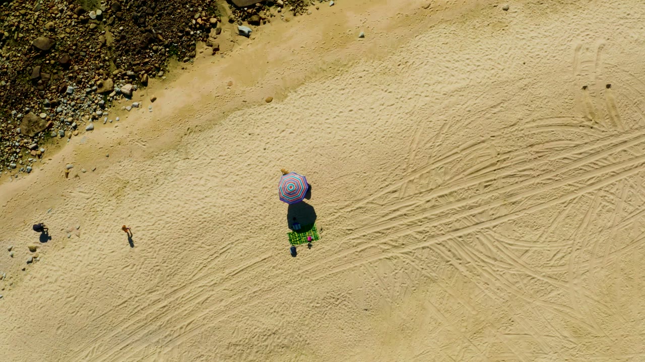 Purple umbrella on open sand with two people near a rocky beach edge.