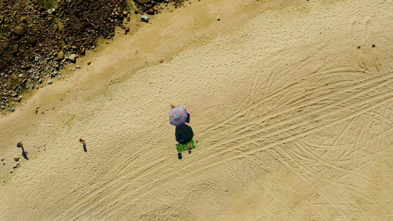 At 0:02, the overhead Chileno Beach view shows more open sand to the right, crossed track marks, the purple umbrella setup, and small visible walkers near the rocks.