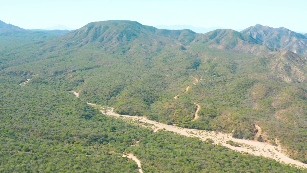 Wide aerial view over El Salado Viejo Los Cabos, Baja California Sur, with a pale dry riverbed crossing green hills and no visible people.