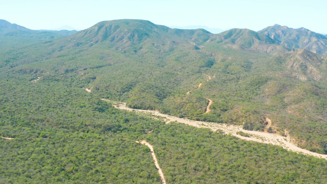 Drone-style view of El Salado Viejo Los Cabos with a curving dirt road and branching pale track running through green brush beneath rocky mountain ridges, with no visible people.