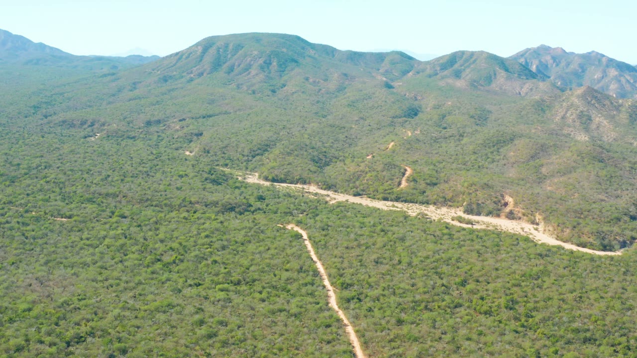 Aerial view of El Salado Viejo Los Cabos with a curving dirt road crossing the green midground and darker mountain slopes behind it, no visible people.