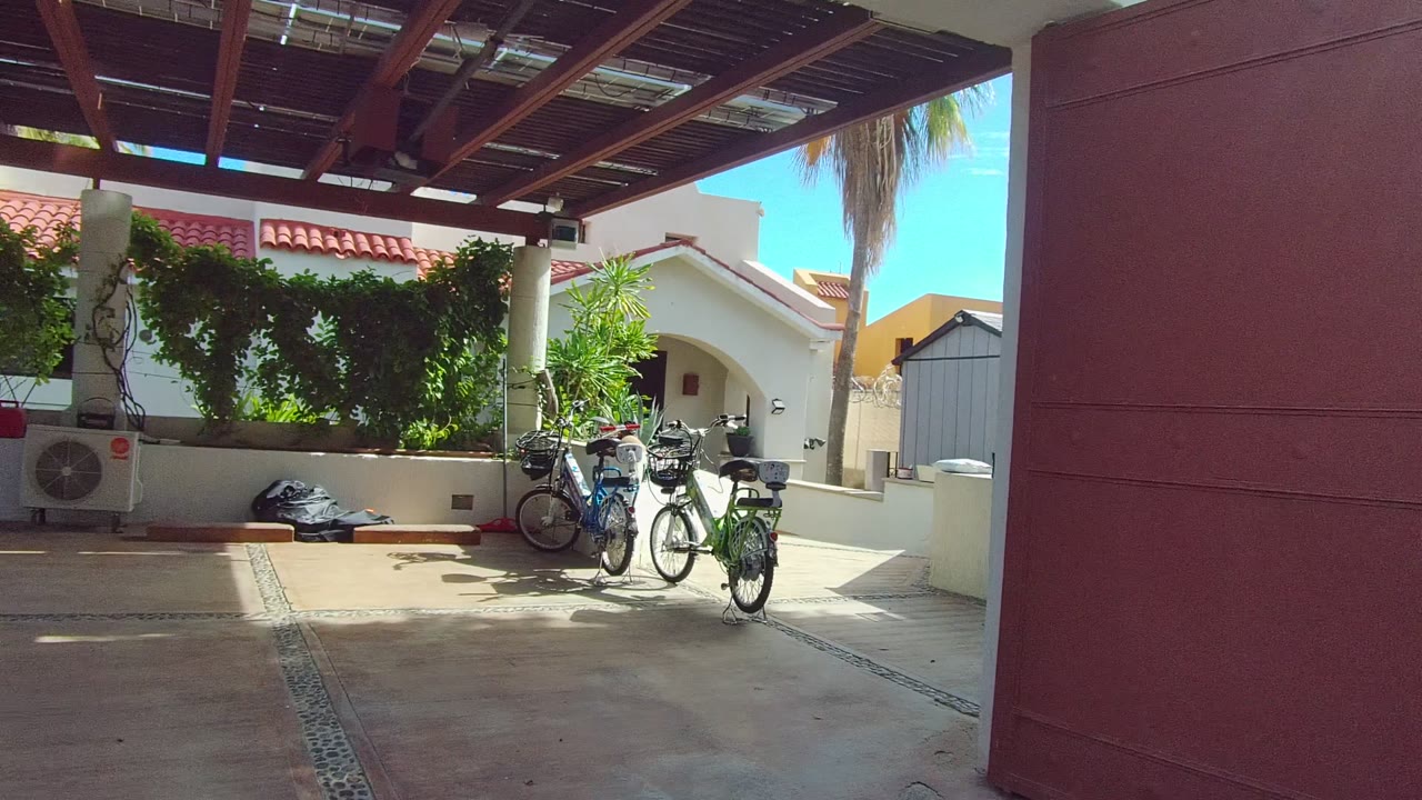 Wide view of an East Cape Los Cabos patio with parked bicycles, a red wall at right, white building exteriors, overhead wooden roof beams, and no visible people.