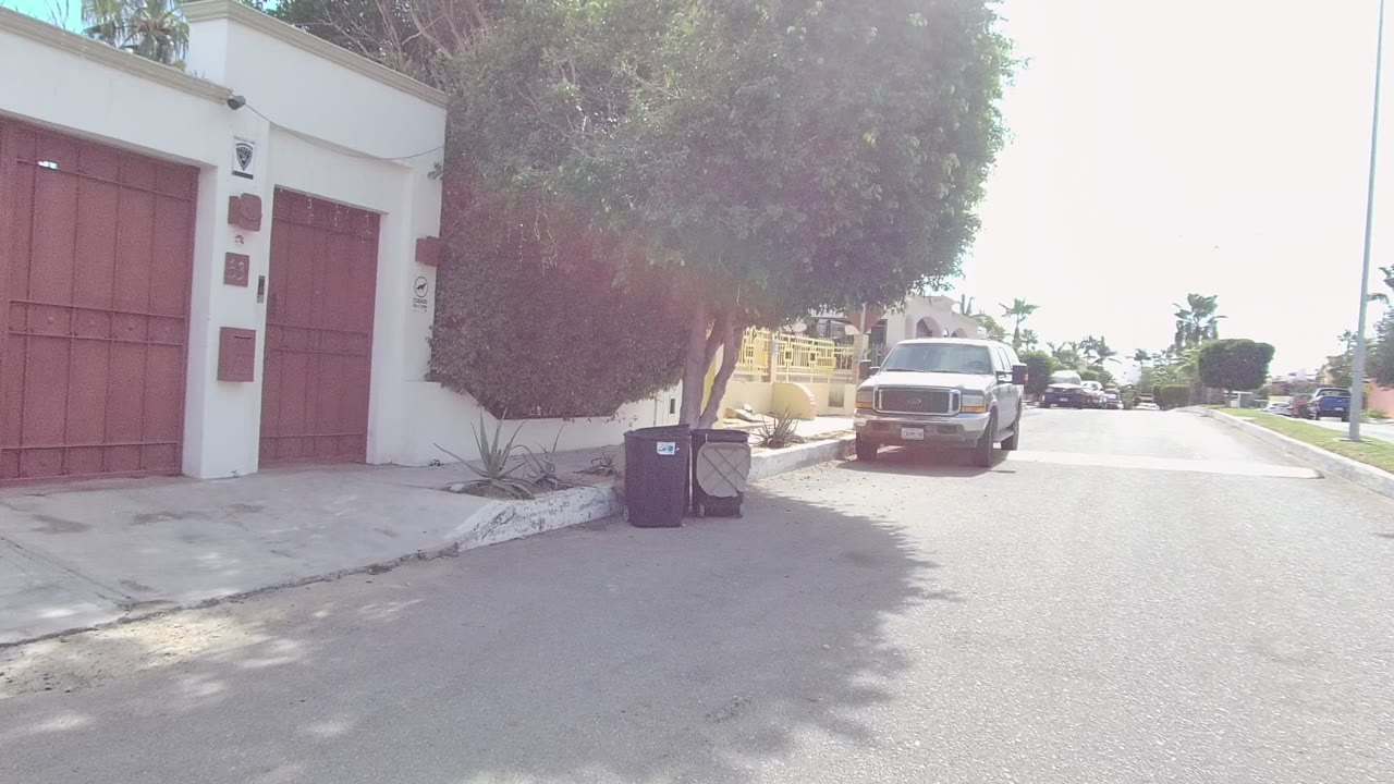 Wide frame in East Cape Los Cabos, Baja California Sur showing red gates on a white residential building, trees, trash bins near the curb, a pickup truck in the street, and no visible people.