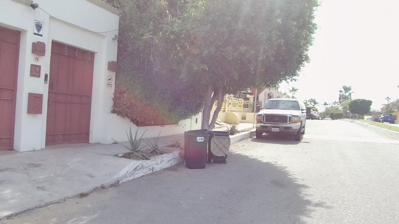 Street-side view of red gates and a white wall with trash bins and a truck farther down the road.
