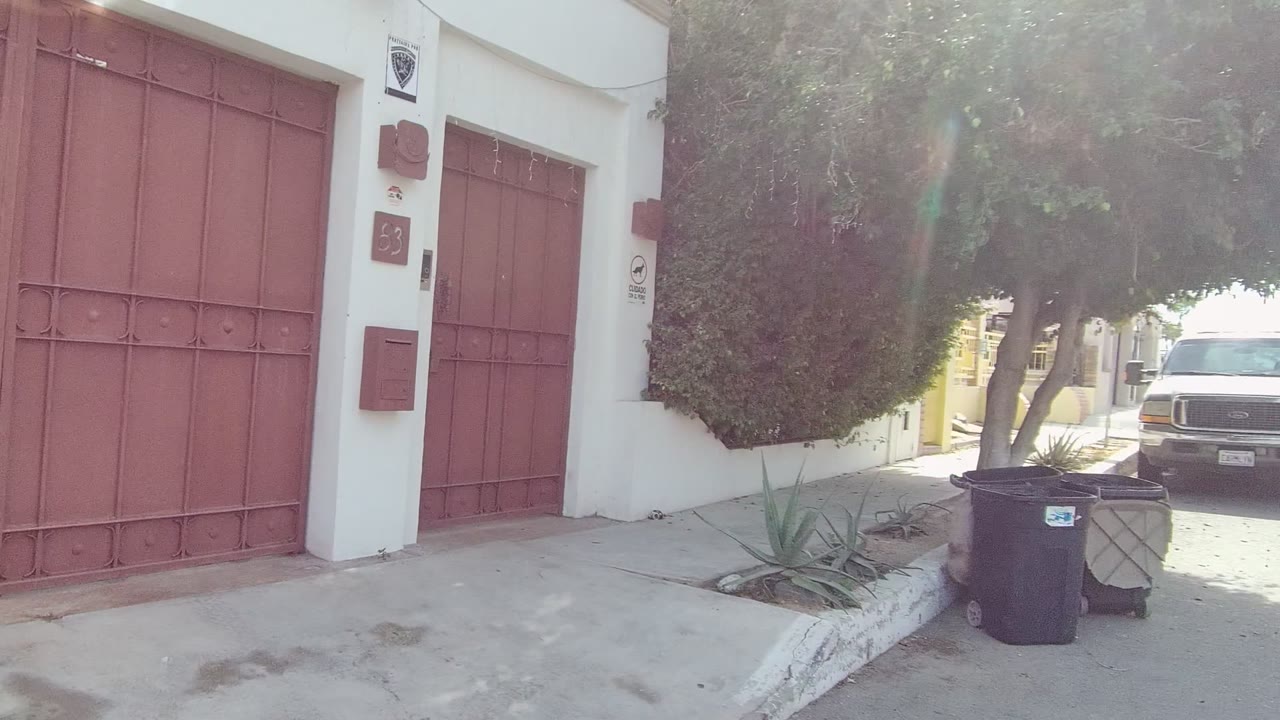 Closer East Cape Los Cabos frame showing tall red metal gates and a smaller red door on a white facade, with trees and trash bins near the curb and no visible people.