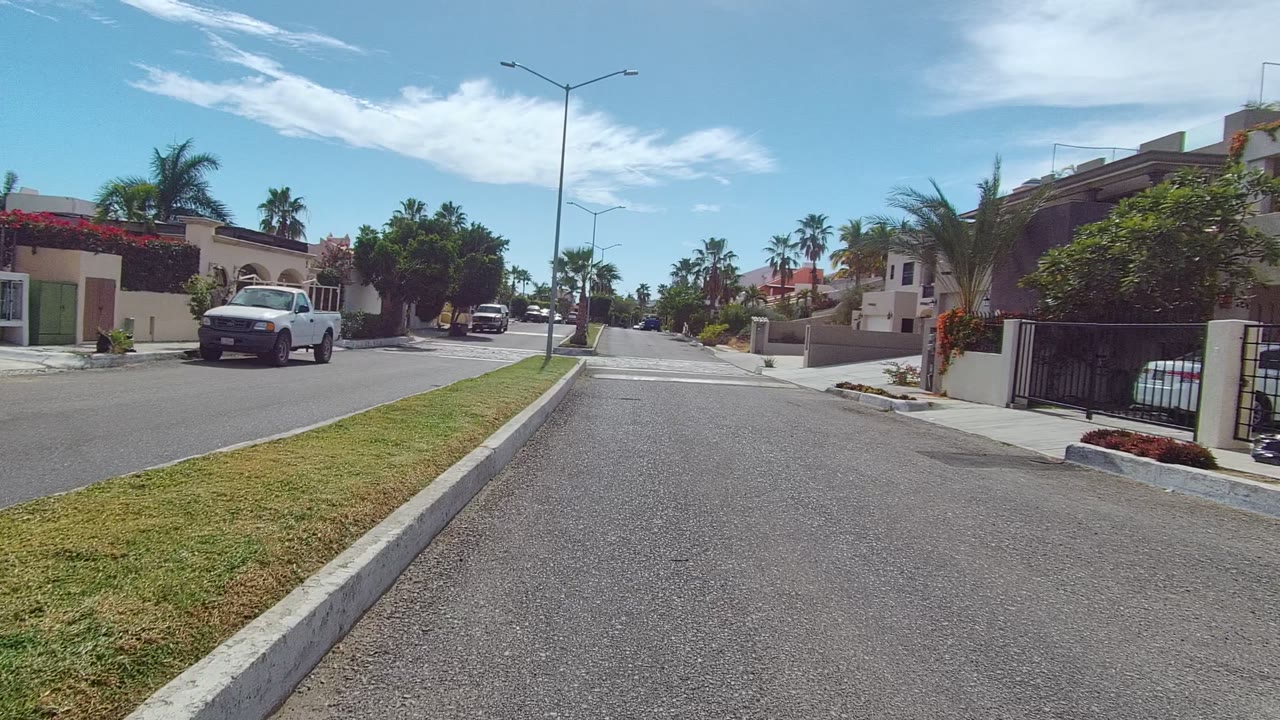 Opening frame of a Los Cabos residential street with a grass median in the foreground, palm trees, walled homes, a white pickup truck at left, blue sky, and no visible people.