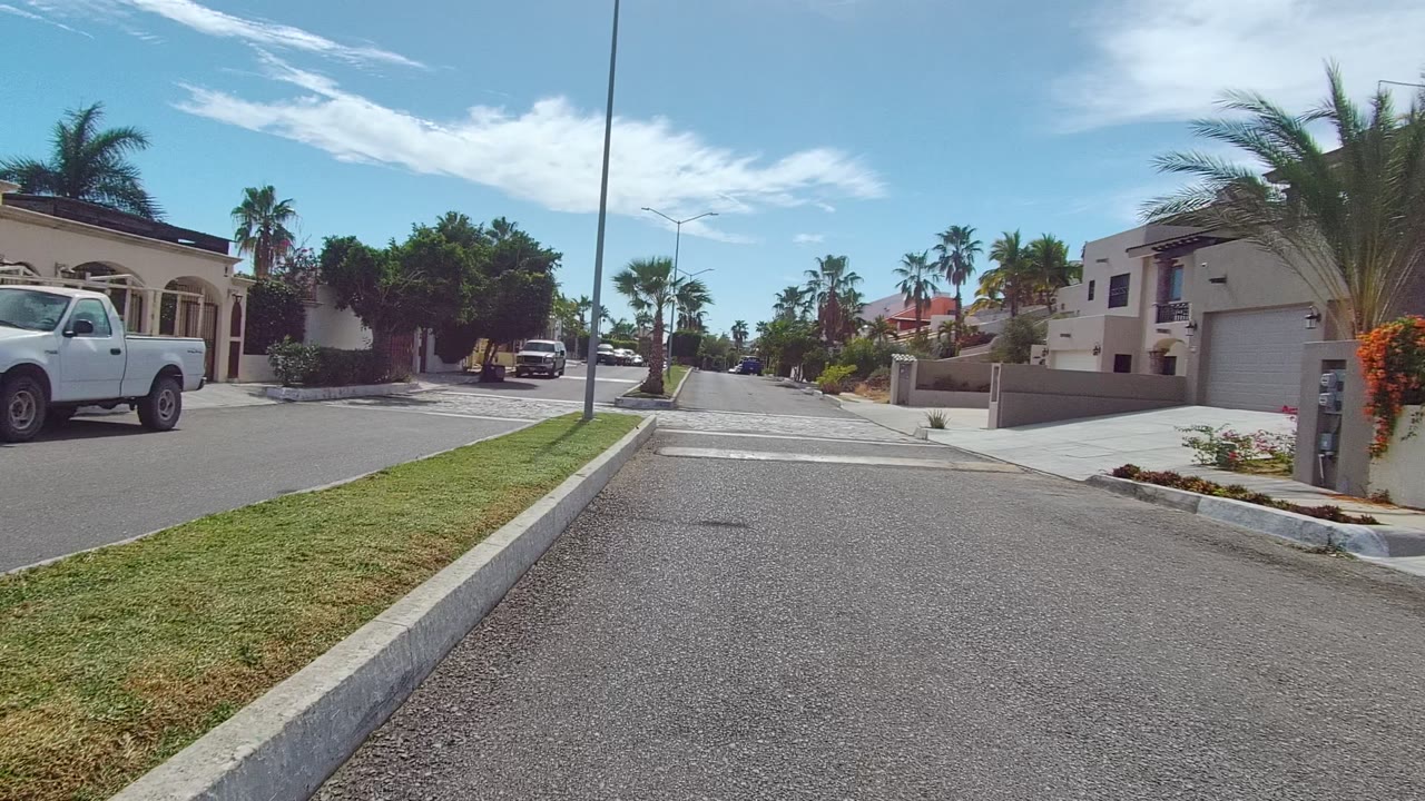 Mid-clip frame in Los Cabos with the road stretching forward, a palm-lined median, a white pickup near a driveway, modern walled homes, blue sky, and no visible people.