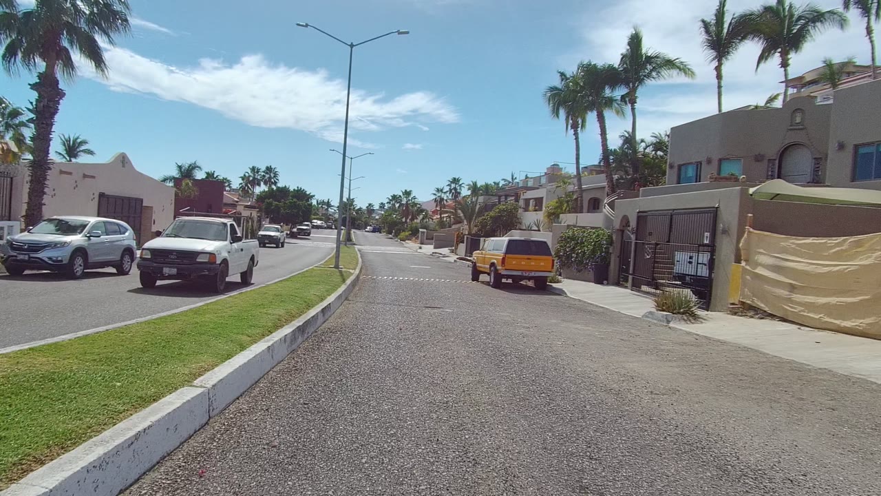 Street-level view in East Cape Los Cabos, Baja California Sur, with a grassy median, low houses, palm trees, white vehicles on the left, a yellow SUV ahead on the right, and no visible people.