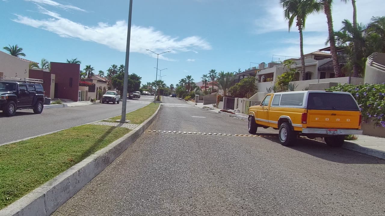 East Cape Los Cabos residential street with the yellow SUV closer on the right, a speed bump across the lane, modern homes, trimmed grass, tall palms, clear blue sky, and no visible people.