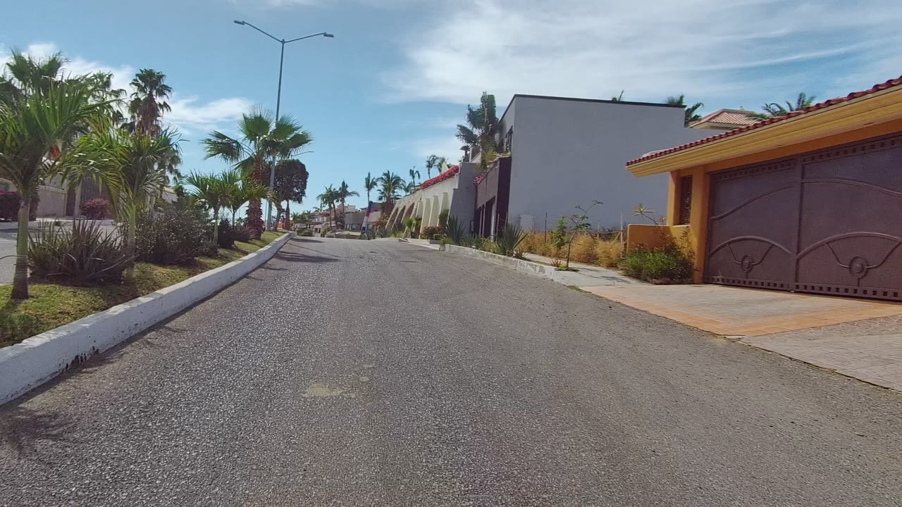 The view advances along a Los Cabos residential street with palms, a driveway, a gated home, low garden planting, and no visible people.