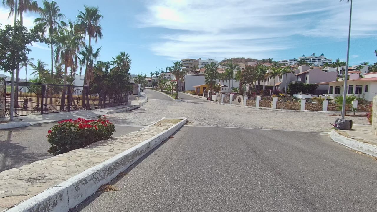 Opening frame in Los Cabos shows a paved approach toward a cobblestone residential street lined with palm trees, white pillars, stone walls, and no visible people.