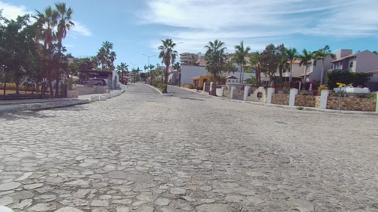 At 0:02, the Los Cabos street view centers on cobblestones, gated homes, white pillars, stone walls, palm crowns, and no visible people under bright daylight.