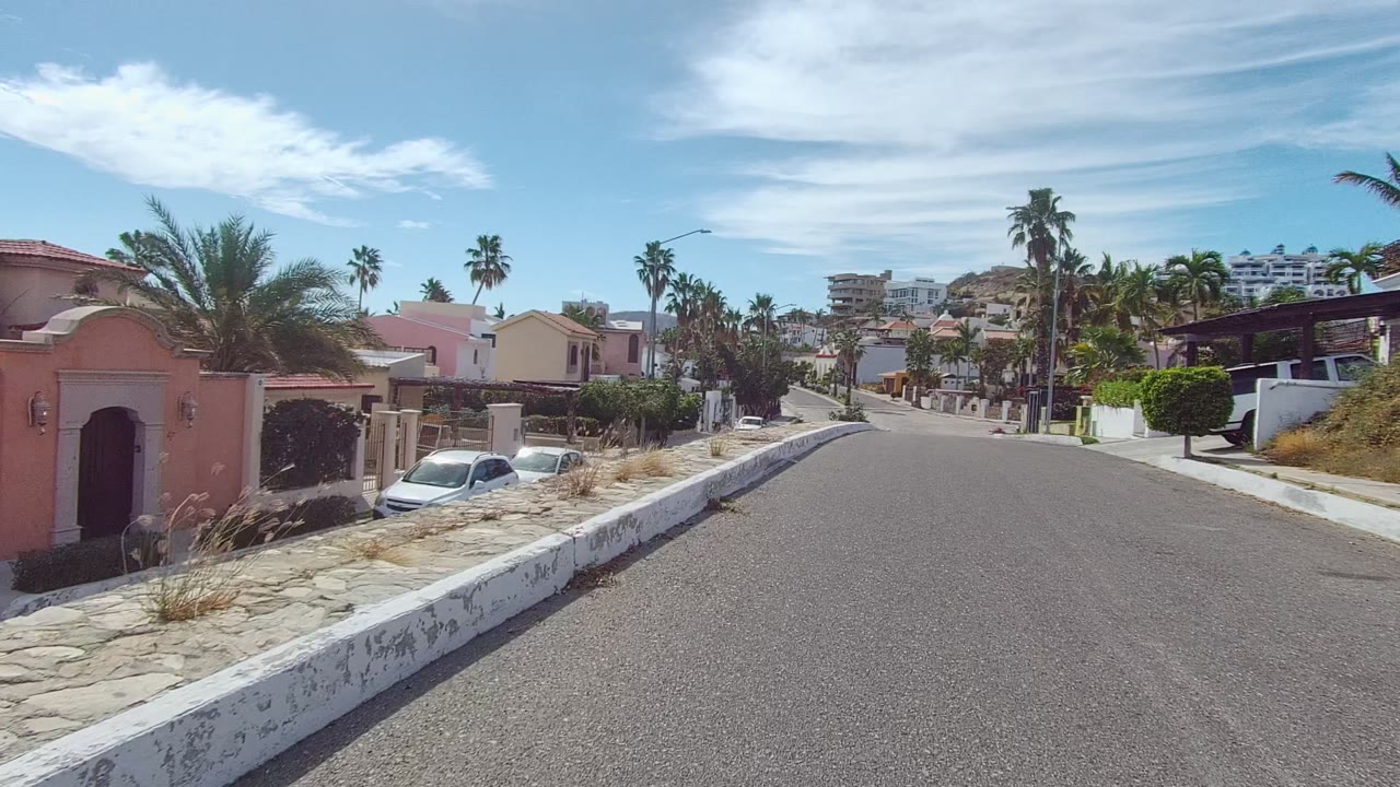 Start of a forward view down a residential street in East Cape Los Cabos, Baja California Sur, with a pink house at left, parked cars, stone sidewalks, palm trees, hillside buildings, scattered clouds, and no visible people.