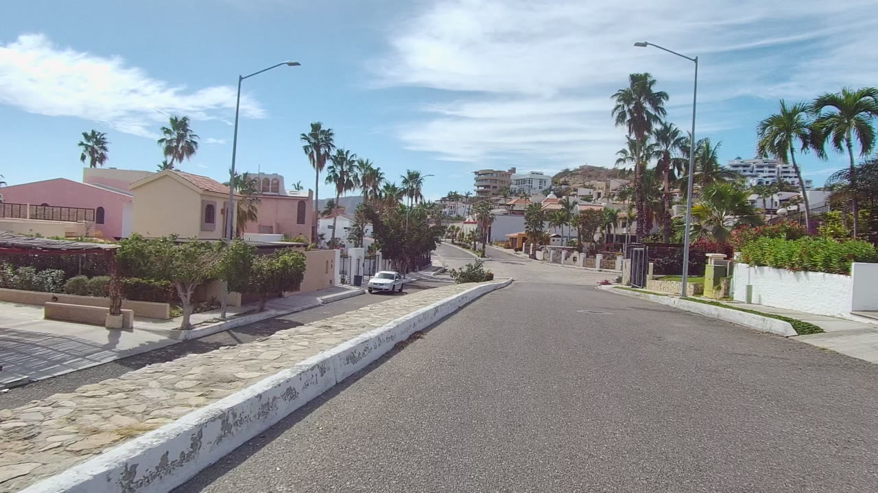 Mid-clip view in East Cape Los Cabos with the camera facing down the residential road past a hedge-lined corner, pale homes, parked cars, palms, stone paving, hillside apartments, and no visible people.