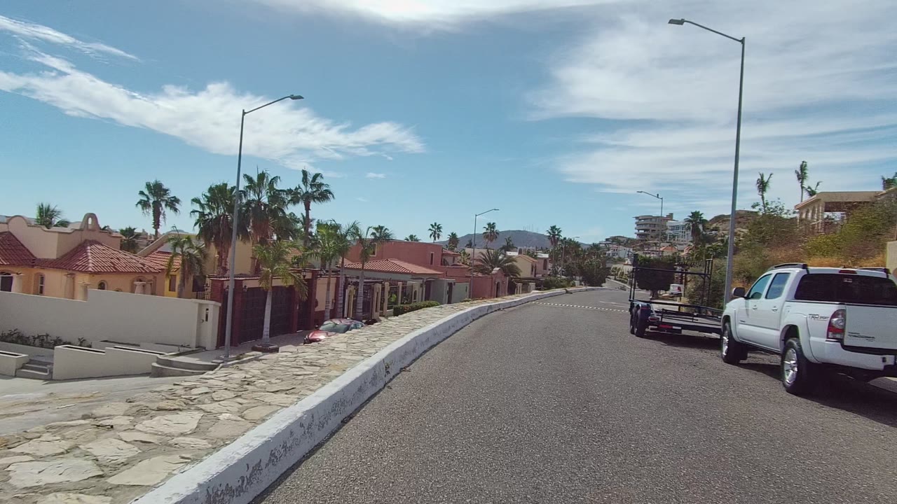Street-level view in Los Cabos, Baja California Sur showing a residential road, red-tiled homes, palm trees, streetlights, parked pickup trucks, and no visible people.