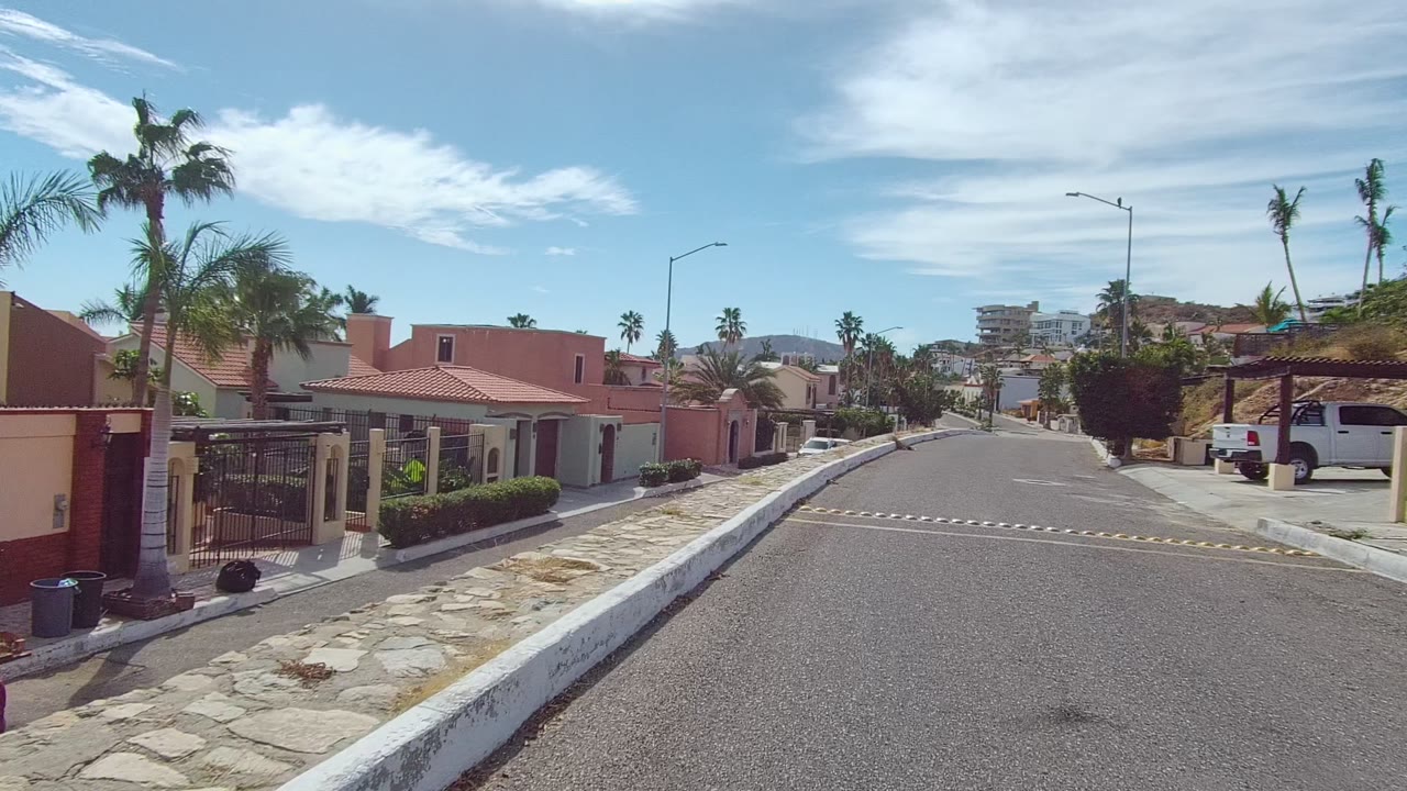 Los Cabos residential corner with a gated red-tile home, a parked utility trailer, palm trees, a white vehicle, a speed bump, and no visible people.