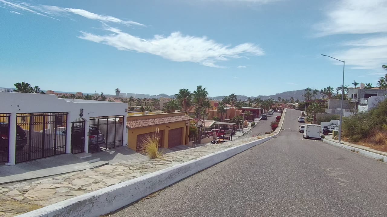 Opening view of a Los Cabos residential street with a white wall at left, a yellow garage, palm trees, parked vehicles, distant hills, and no visible people.