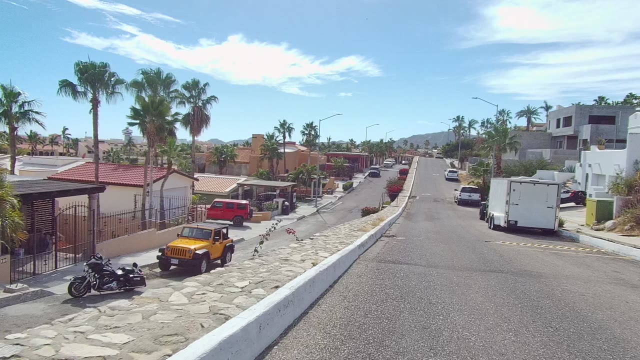 Mid-clip Los Cabos frame with a gated house at right, a motorcycle near the curb, a yellow Jeep, a white trailer across the street, palm trees, and no visible people.