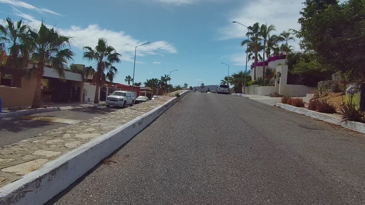 Mid-clip frame in Los Cabos shows the uphill asphalt road beside a stone wall, parked vehicles, palm trees, residential gates, distant low hills, and no visible people.