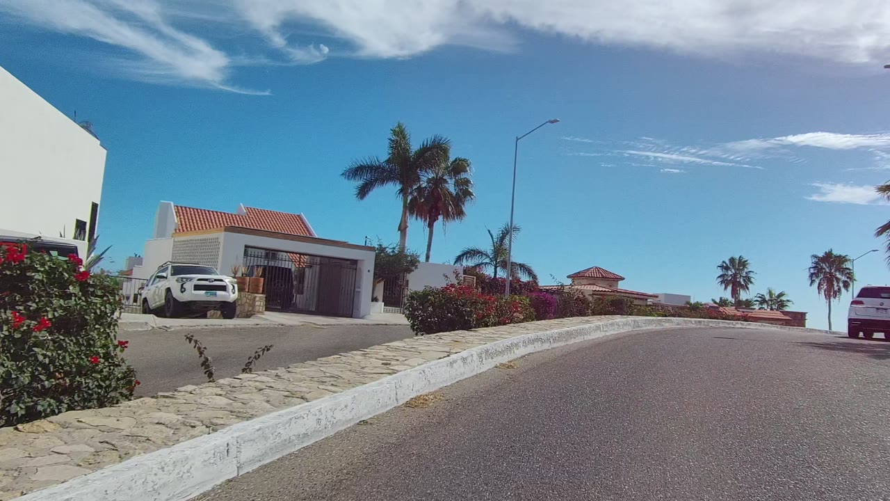 Opening frame of an East Cape Los Cabos residential street with a white curb, stone sidewalk edge, palm trees, flowering shrubs, a parked white car, red-tiled roofs, blue sky, and no visible people.
