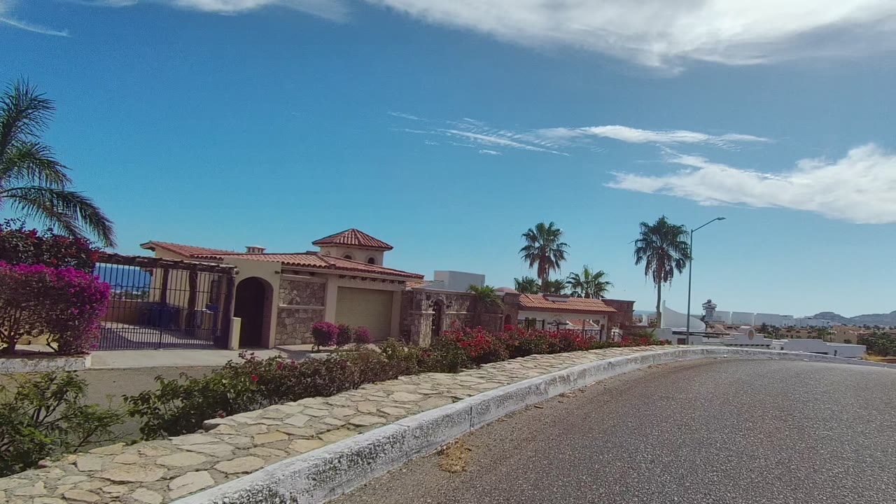 East Cape Los Cabos frame with a gated red-tile residence on the left, palms over the wall, flowering plants along the curb, the road curving ahead, distant hills, and no visible people.