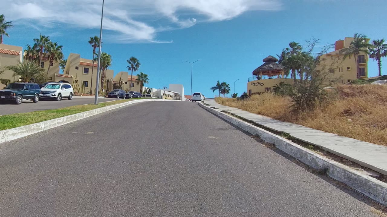 Street-level view of a Los Cabos residential road with tan houses, parked vehicles, palm trees, sidewalks, and no visible people at the start of the clip.