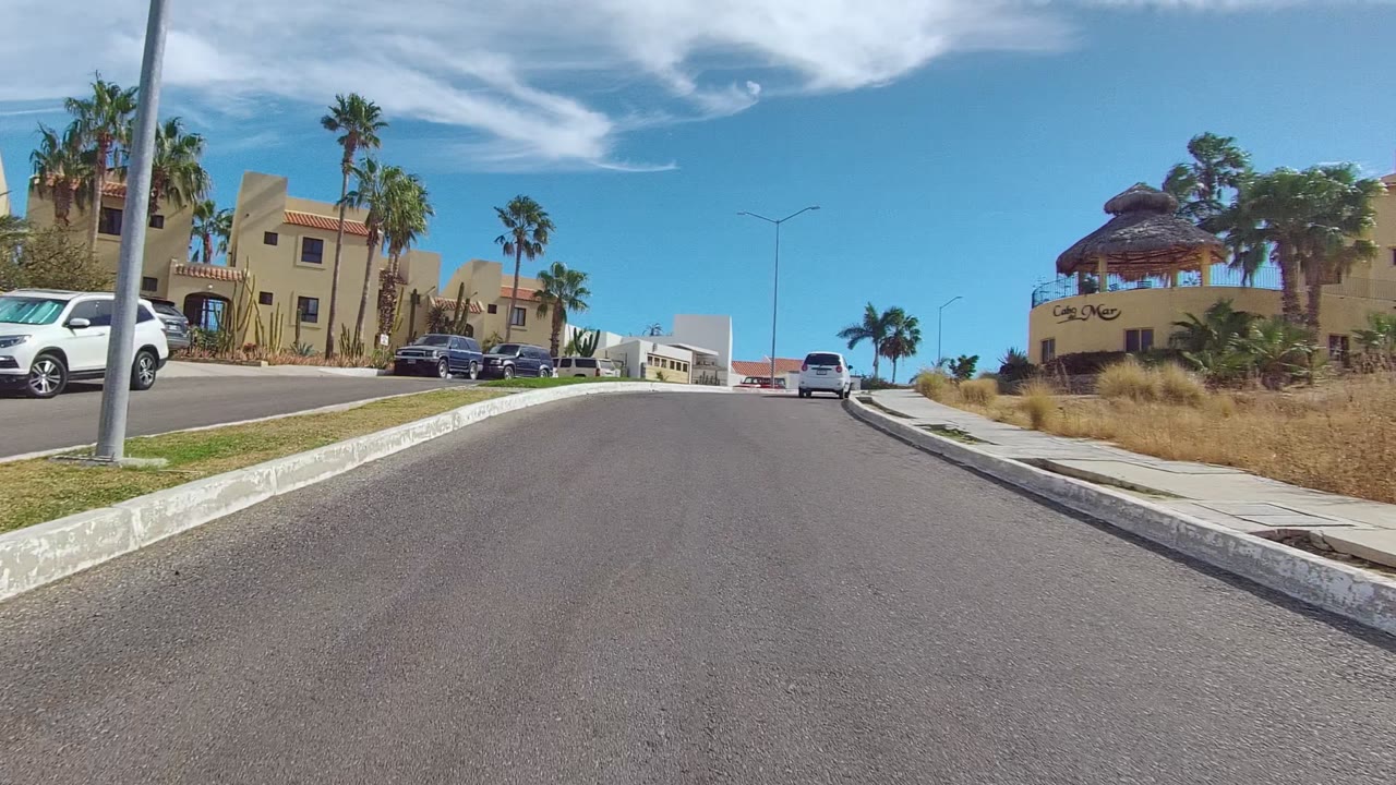 Road climbing past tan homes, palms, parked cars, sidewalk, and curb.