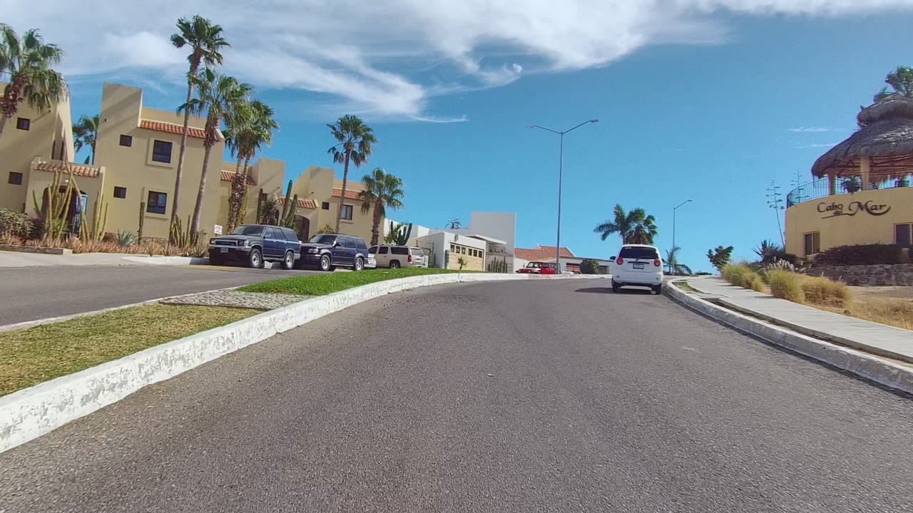 Los Cabos neighborhood street view passing a tan corner building, parked pickup trucks, palm trees, a sidewalk, and no visible people.