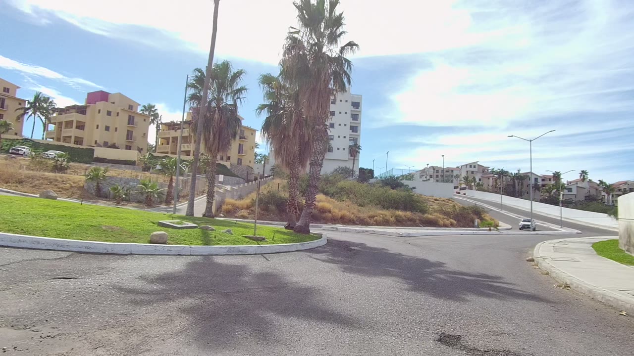 Opening view of a curving residential road in Los Cabos with tall palms, a green lawn, dry roadside grasses, tan and white buildings, and no visible people.