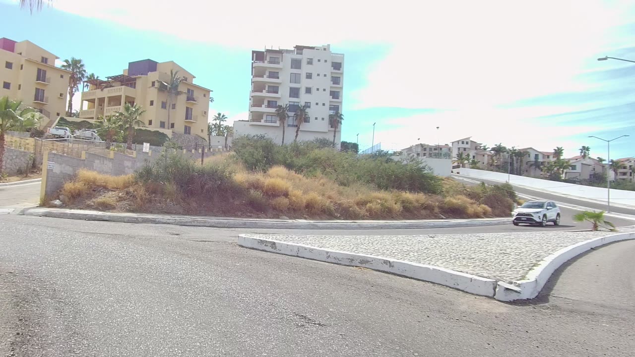 The road opens toward a pale curb and planted island in Los Cabos, with a white car at the right edge, dry grass, palms, apartment buildings, and no visible people.