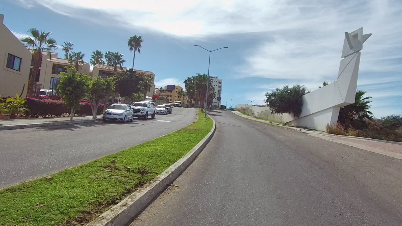 Street-level view in Los Cabos, Baja California Sur, with parked cars along a residential road, palm trees, beige buildings, a white angular roadside structure, and no visible people.