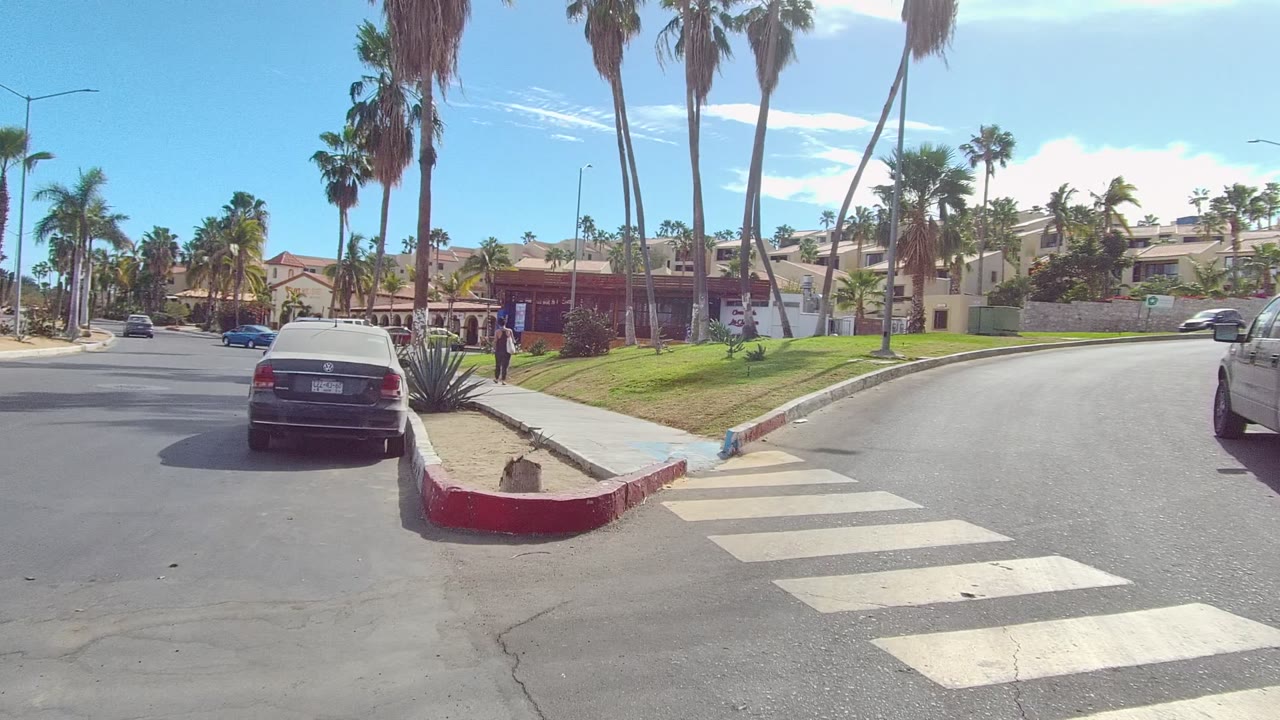 Opening frame of a Los Cabos residential street with a crosswalk in the foreground, red curb, parked car, sidewalk, palms, grass, and no visible people.