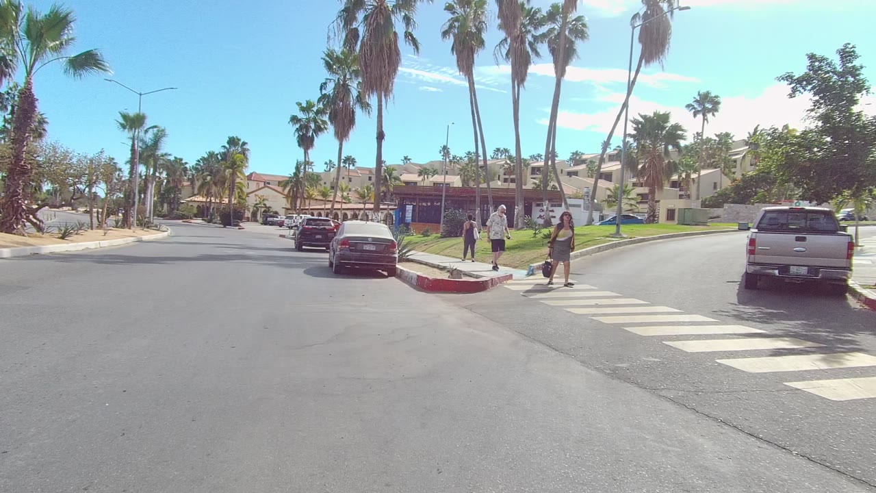 At 0:00 in Los Cabos, Baja California Sur, a wide sunny intersection shows a marked crosswalk, parked and moving cars, palm trees, and two pedestrians near the curb.
