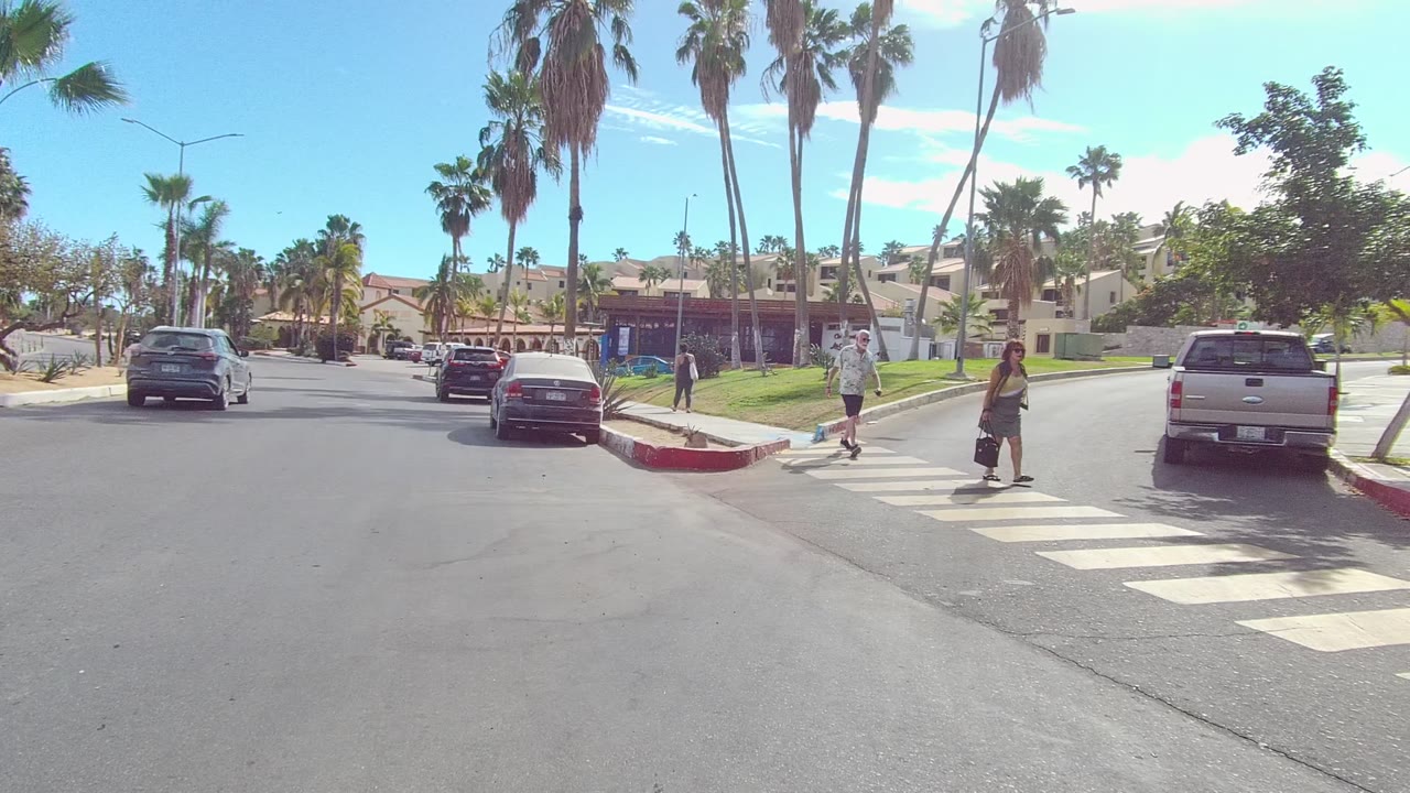 At 0:02 in Los Cabos, Baja California Sur, two adults are visible on the crosswalk near the right side of frame, with a pickup, parked cars, palms, and residential buildings behind them.