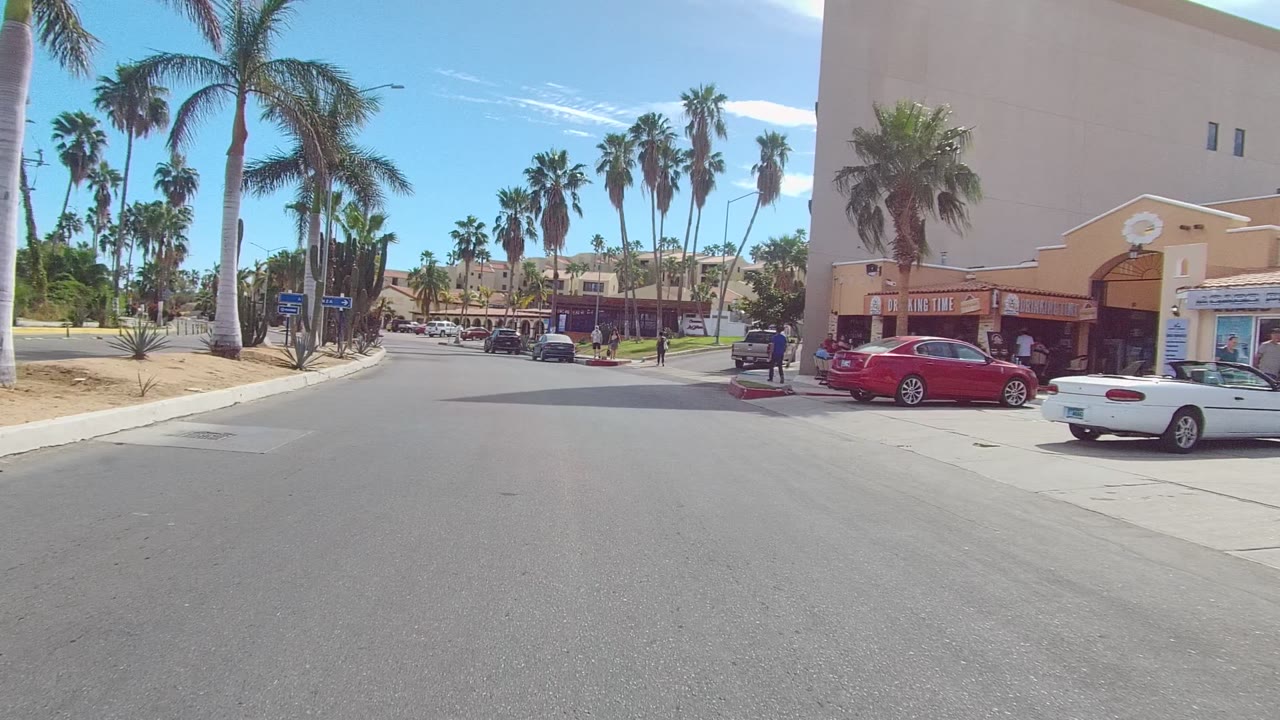 Opening view of an East Cape Los Cabos street with tall palms, a storefront corner, parked cars, cactus landscaping, and people visible near the right sidewalk.