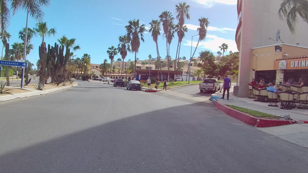 Street-level frame in East Cape Los Cabos showing a storefront patio at right, a red car by the curb, tall palms overhead, and people visible on the sidewalk.