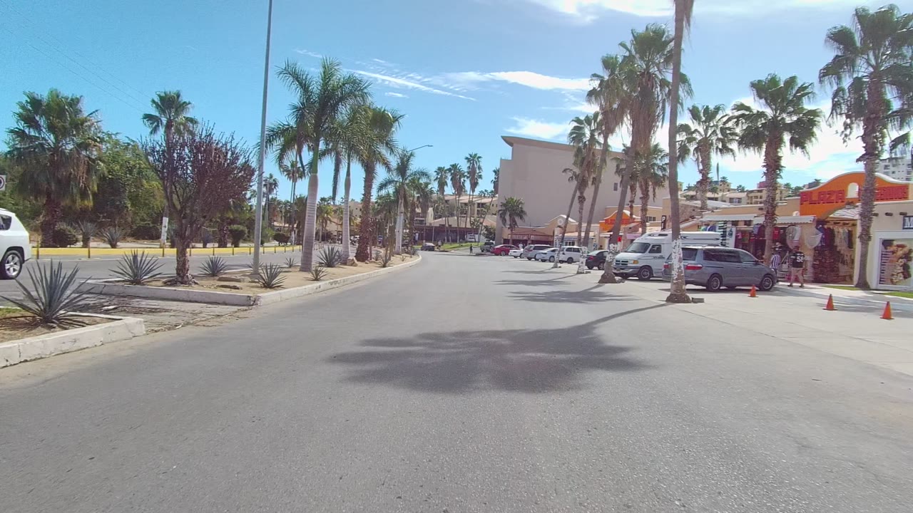 Opening view of a Los Cabos street with palms, desert landscaping, parked cars, orange awnings, blue sky, and no visible people.